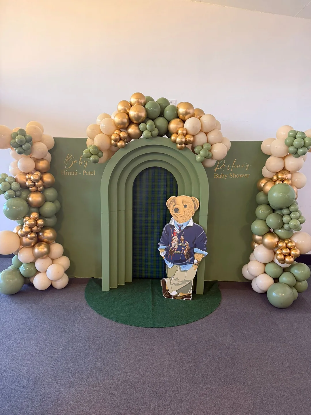 A baby shower with green arched backdrops, a sage and gold garland and a teddy bear cutout