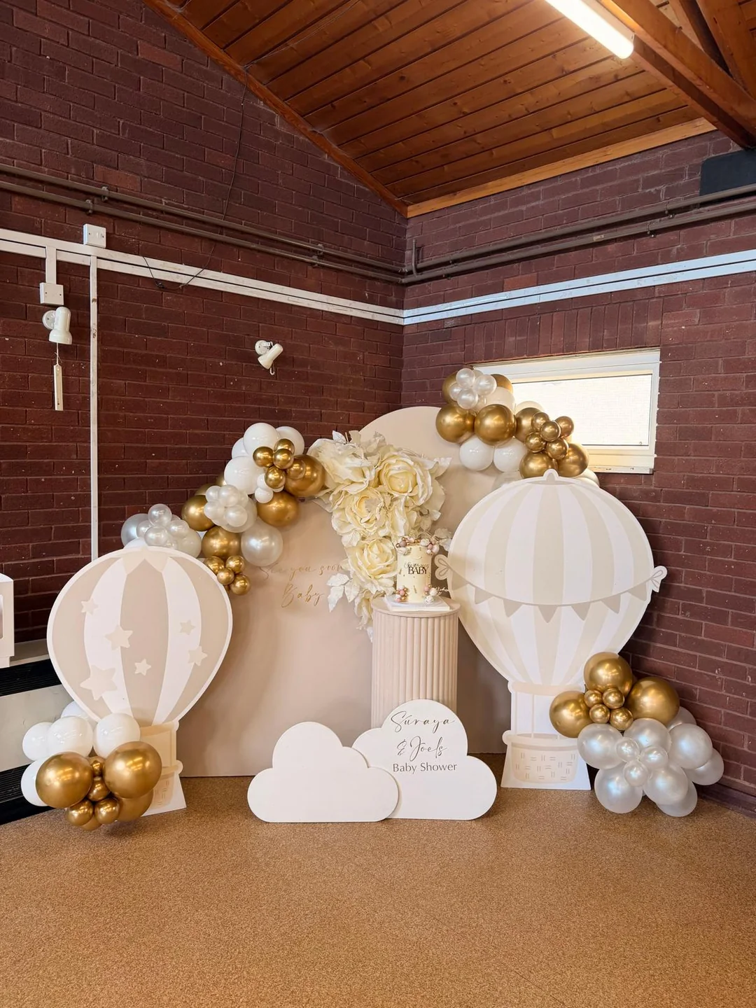 A baby shower backdrop with hot air balloon props, clouds and a white and gold balloon garland