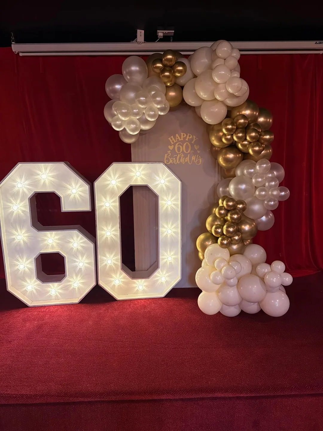 A light up 60 with a white and gold balloon garland against a red drape