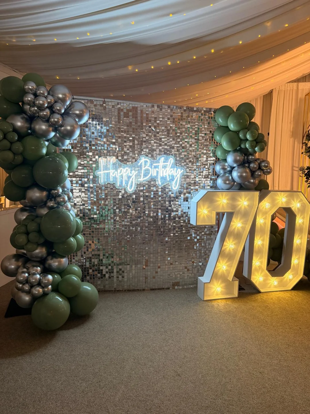 A silver sequin wall with a happy birthday neon sign, a green and chrome garland and a light up 70