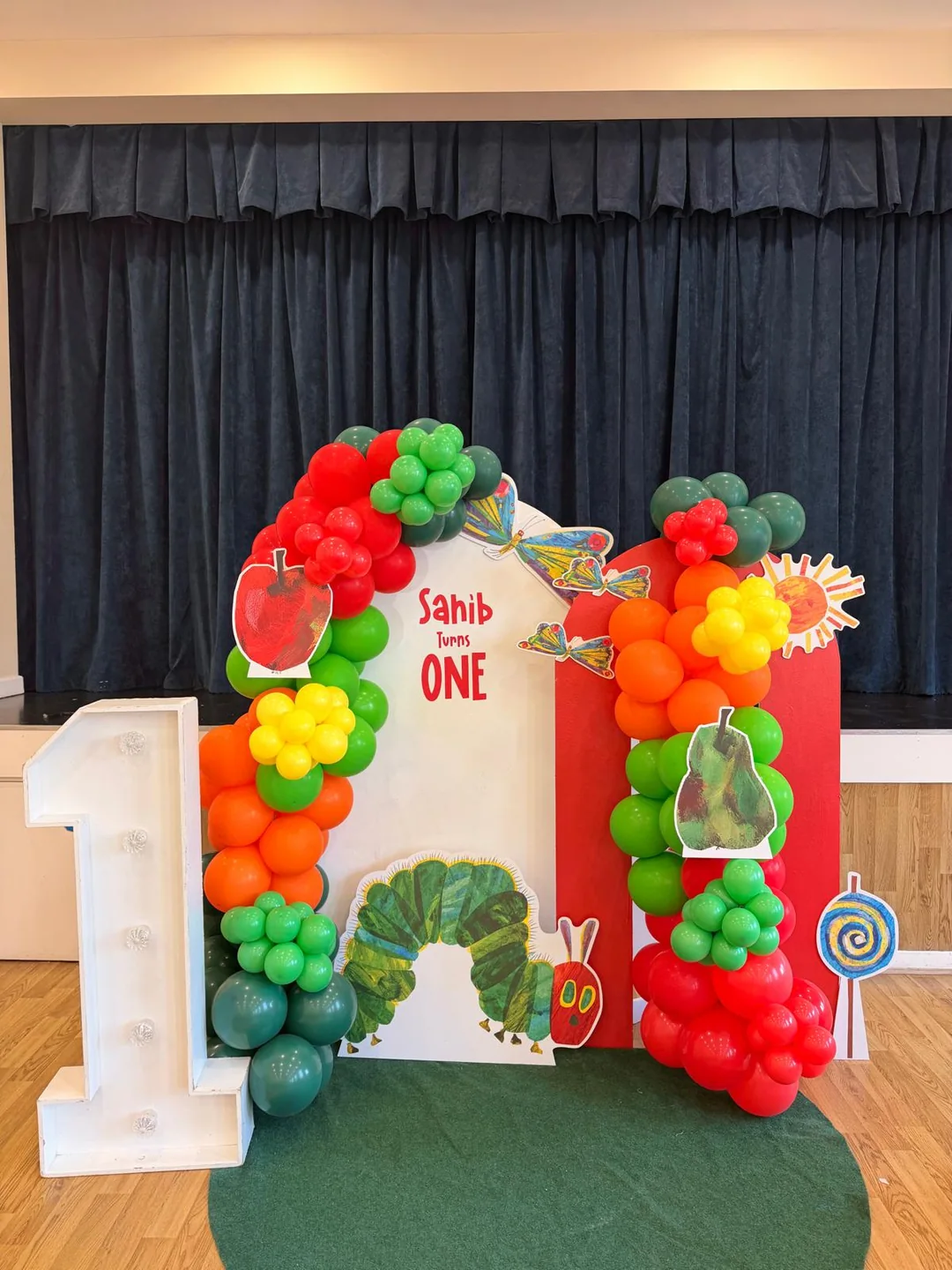 A Very Hungry Caterpillar first birthday with a red, green and orange balloon arch and a light up number one