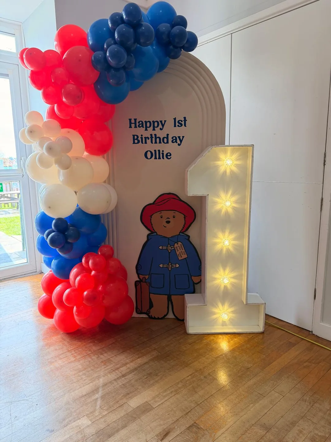 A Paddington first birthday with a red, blue and cream balloon garland and a light up number one