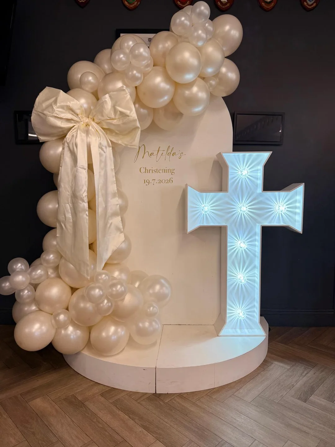 A christening display with a cream and pearl balloon garland, a large bow and an illuminated cross