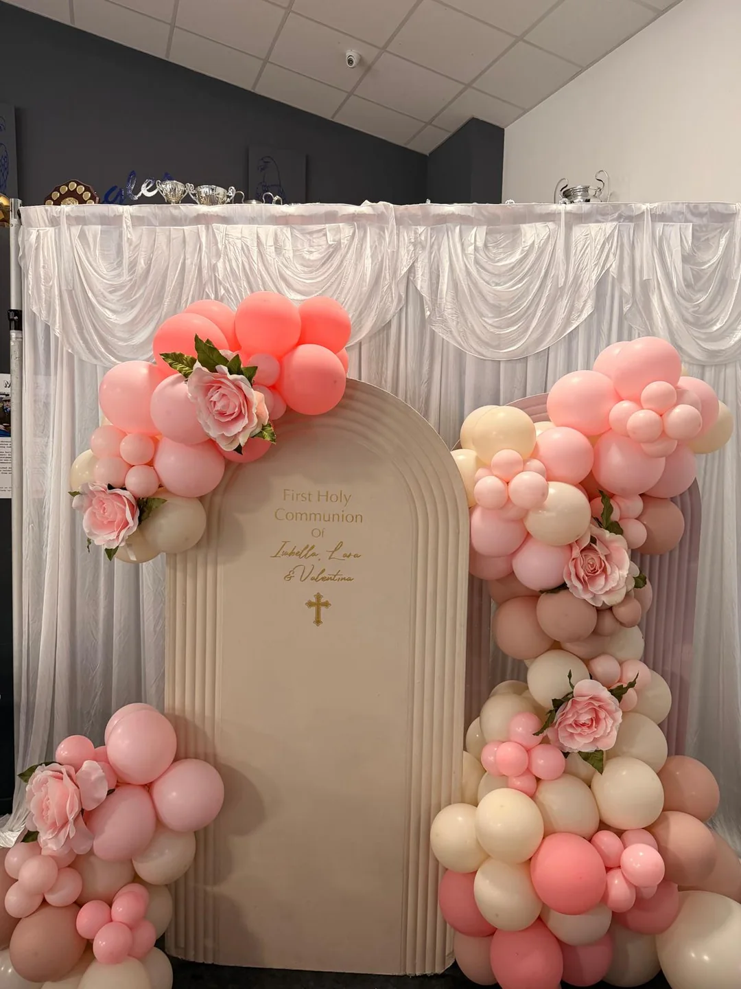 A holy communion backdrop with a blush and cream balloon garland, flowers and a gold cross