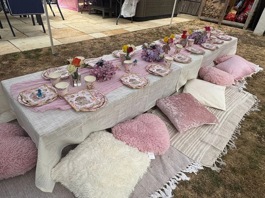A long garden table laid with a pink runner, fairy themed plates and cups, flowers and floor cushions