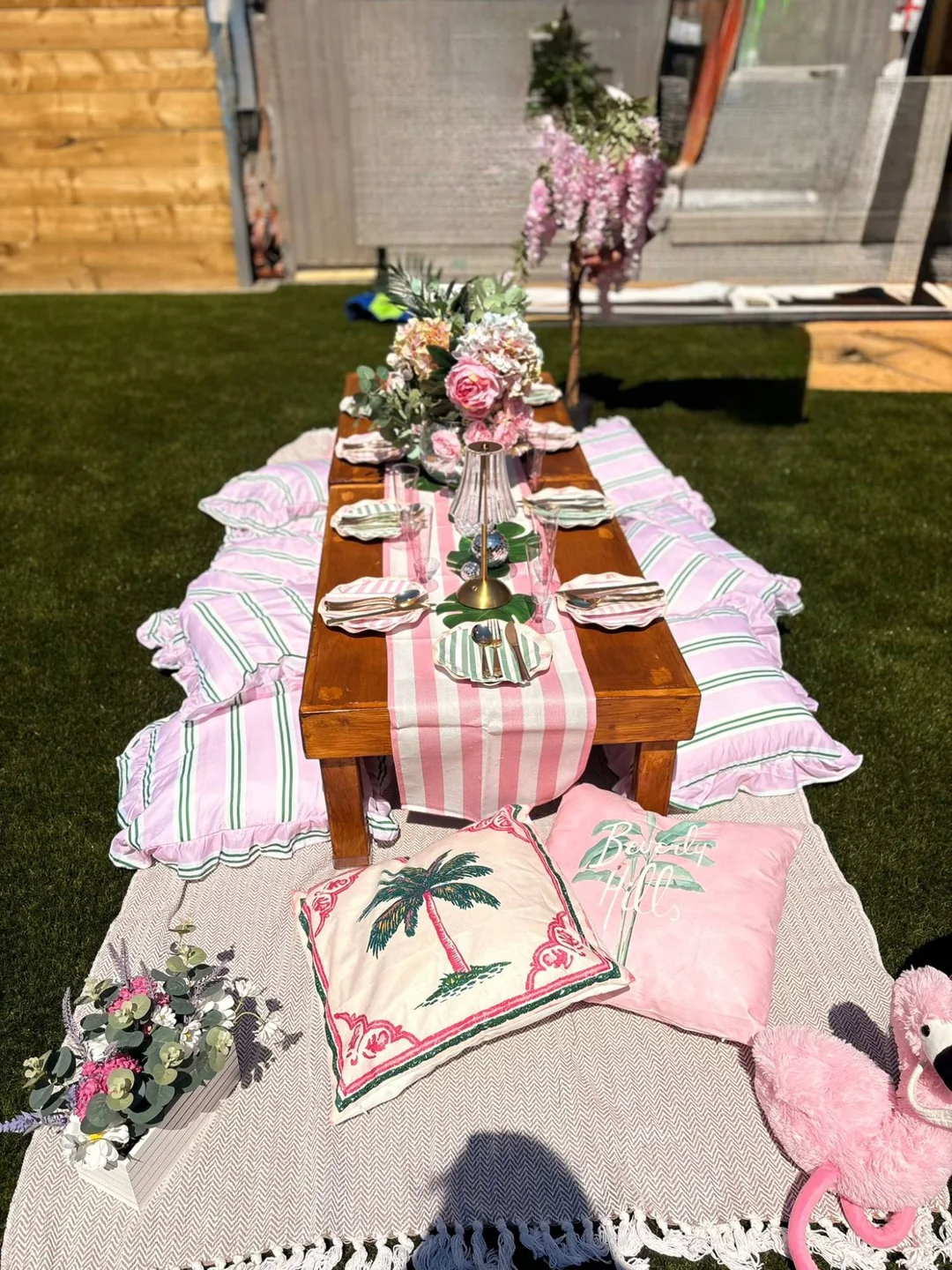 A low wooden table on the grass laid with a pink striped runner, scalloped plates and ruffled floor cushions