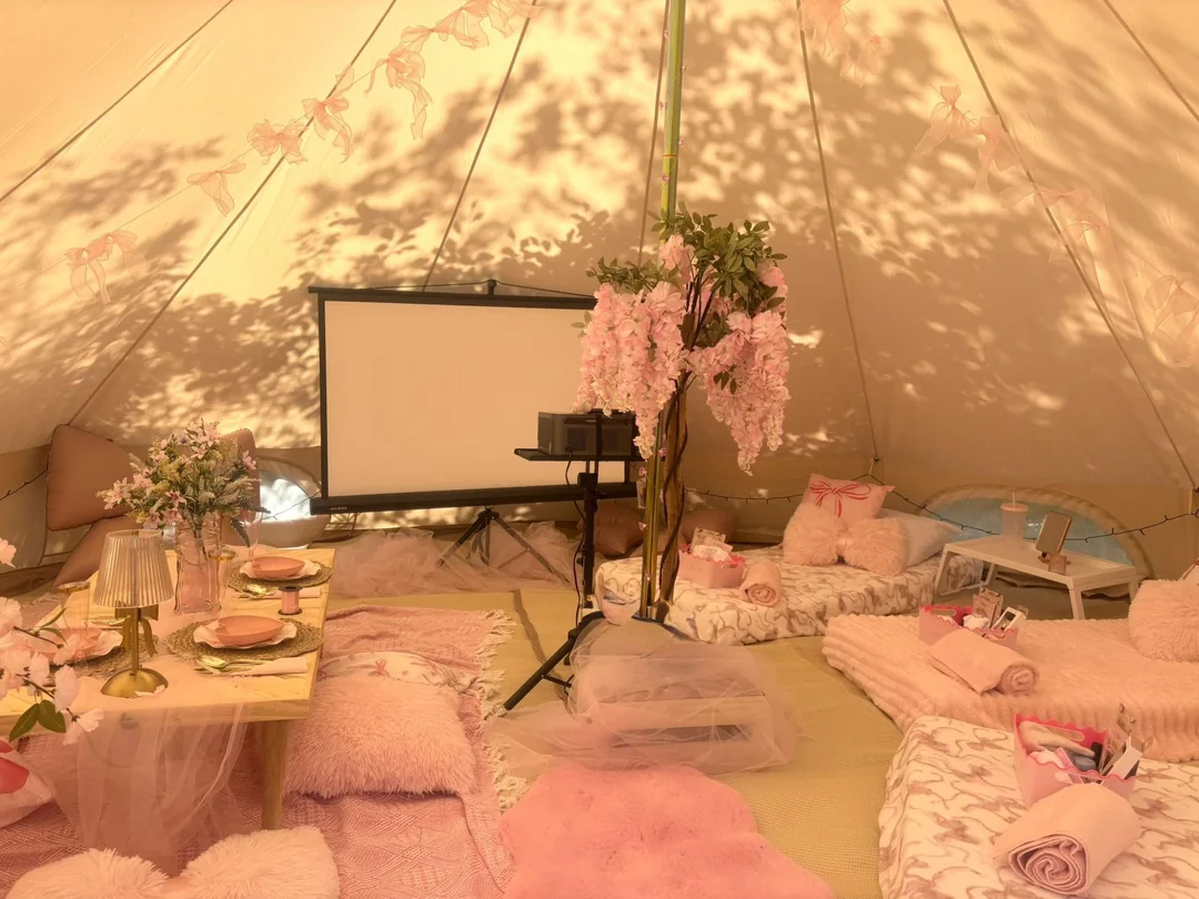 The inside of a bell tent set up for a sleepover, with floor mattresses, pink throws, a low table laid for tea and a projector screen
