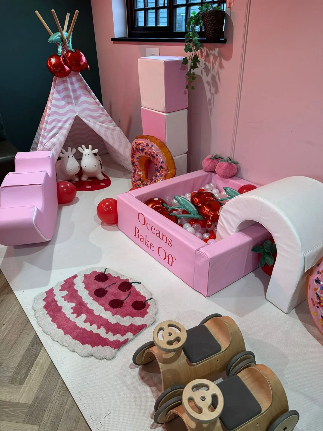 A pink ball pit printed with a child's name, filled with cherry balloons, beside a striped teepee and a giant doughnut balloon