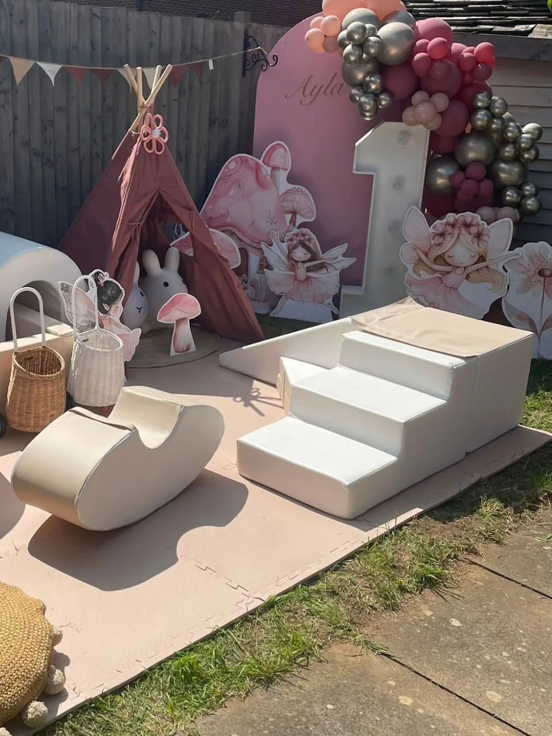 Cream soft play with steps, a rocker and a play house set up on pink matting in a garden, beside a teepee and a fairy backdrop