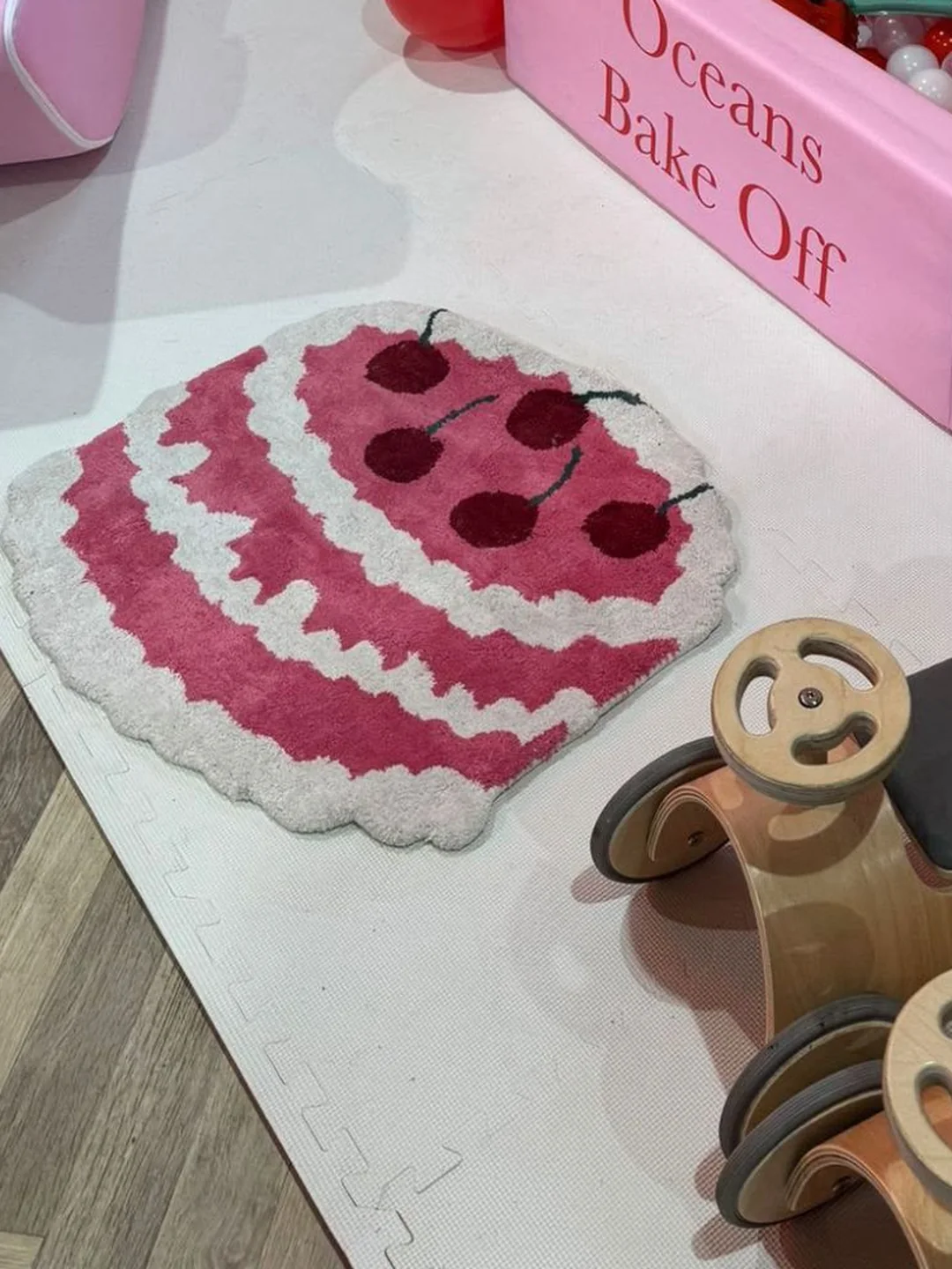 A cherry cake rug and wooden ride on cars on foam matting next to a pink ball pit