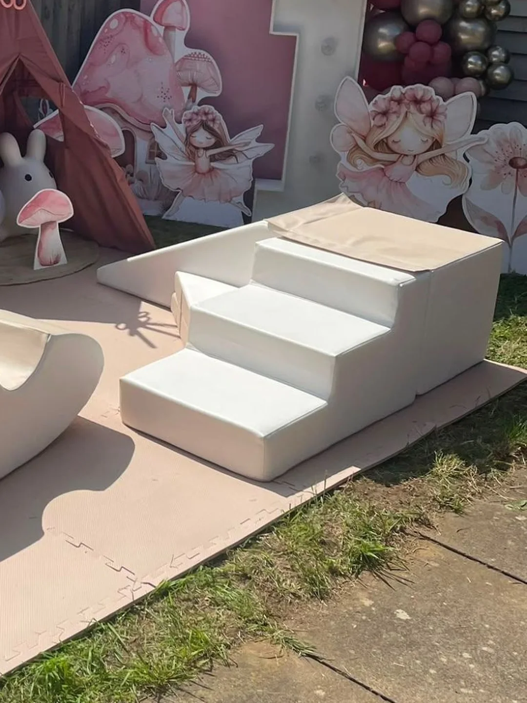 Cream soft play steps on pink matting in a garden, under a pink and chrome balloon garland