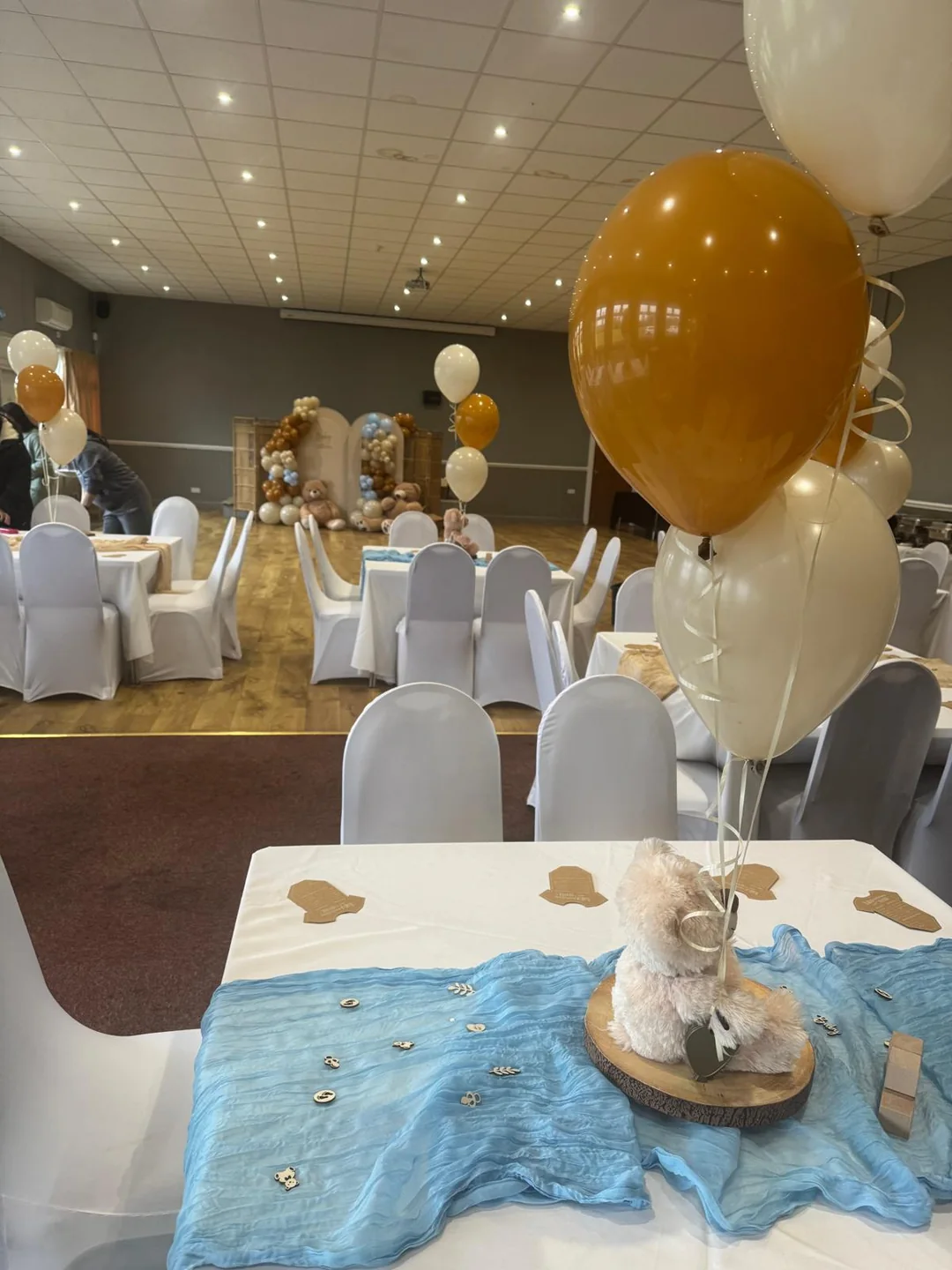 A hall laid with covered chairs, round tables, teddy centrepieces and balloon bouquets