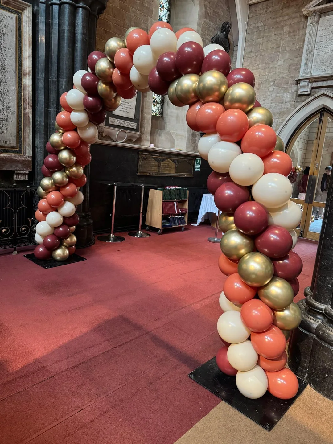 A burgundy, coral, gold and cream balloon arch across a grand brick hall