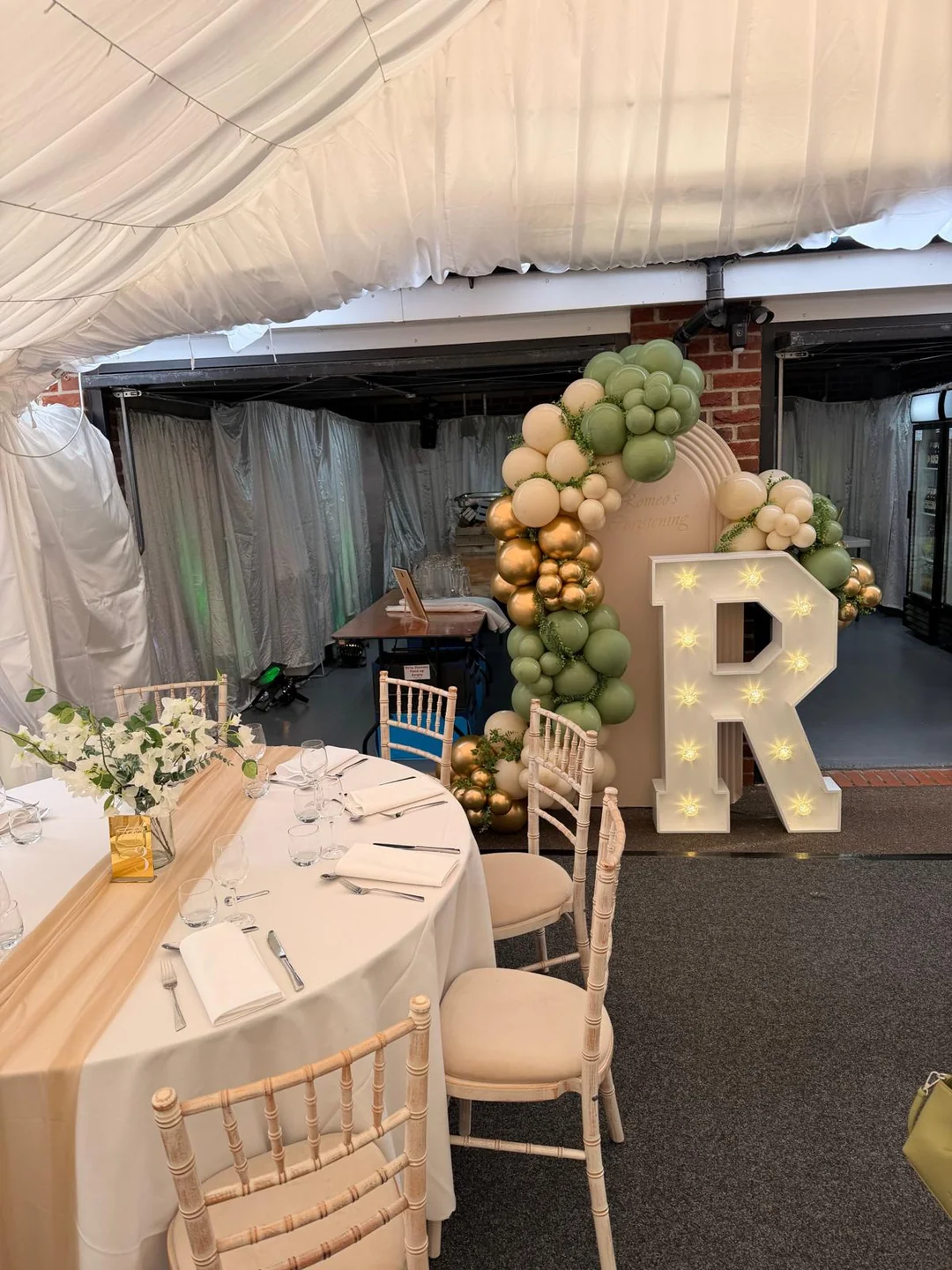 A marquee dressed with a sage and gold balloon garland, a light up letter and a laid round table